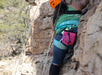 Person climbing a rock wall with safety gear and a pink bag.