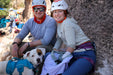 Two climbers with a dog sitting on rocks, wearing helmets and sunglasses.
