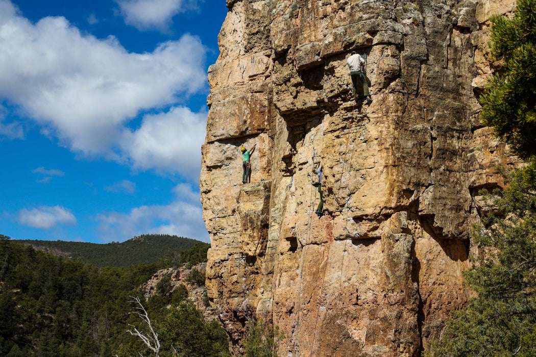 Person climbing a rocky cliff face with a blue sky and green trees in the background