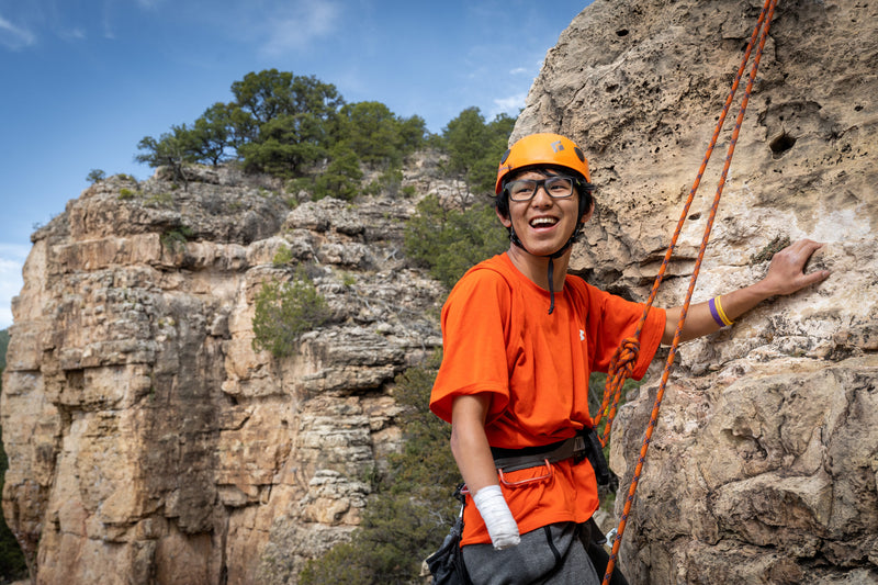 Person in orange shirt and helmet climbing a rock face with a clear sky and trees in the background.