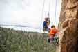 Two climbers on a rock face with a scenic background of trees and sky.
