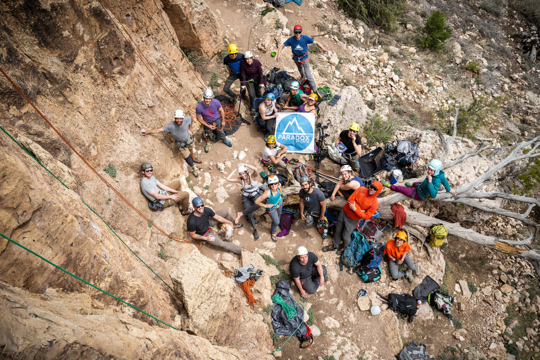 Group of people gathered on a rocky mountain ledge with a Paradox Sports logo in the center.
