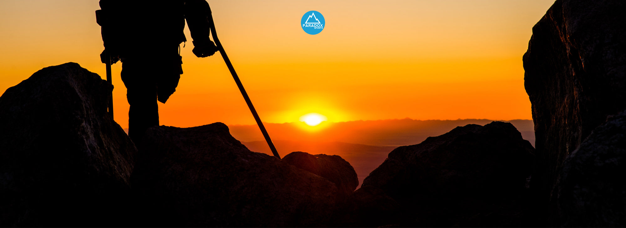 A silhouette of an amputee climber at the peak of a mountain as the sun sets behind the horizon.