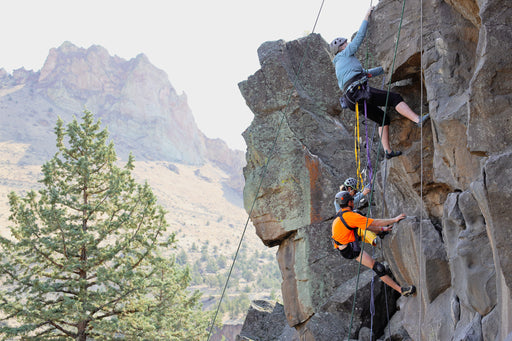 Two climbers on a rocky cliff with mountains in the background.
