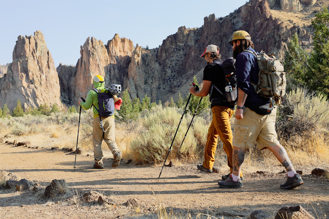Three hikers with backpacks and hiking poles walking on a trail with rocky cliffs in the background.