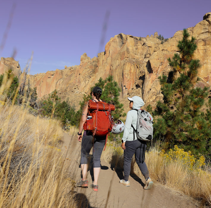 Two hikers with backpacks walking on a trail with rocky cliffs and trees in the background.