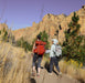 Two hikers with backpacks walking on a trail with rocky cliffs and trees in the background.