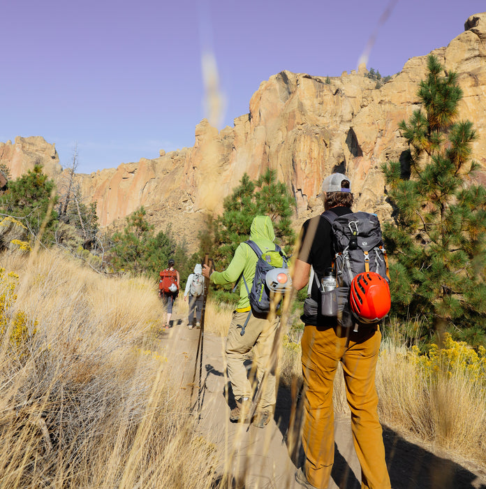 Hikers on a trail with rocky cliffs and trees in the background.
