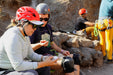 Two people sitting on rocks wearing helmets and sunglasses, with a third person in the background.