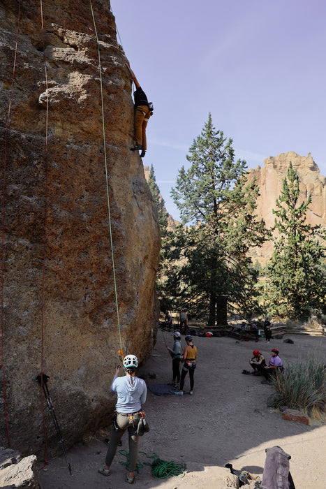 Person rock climbing on a large rock face with others in a desert setting.
