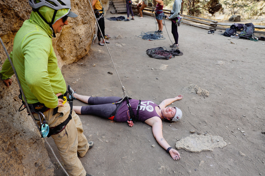 Person in a purple shirt lying on the ground with climbing equipment around, while another person in a green jacket observes.