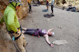 Person in a purple shirt lying on the ground with climbing equipment around, while another person in a green jacket observes.