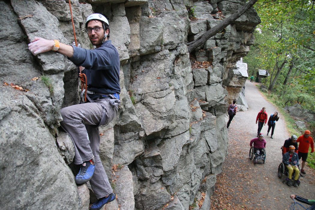 Person climbing a rocky cliff with others walking on a path below.