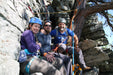 Three climbers taking a break on a rocky mountain ledge with climbing gear.