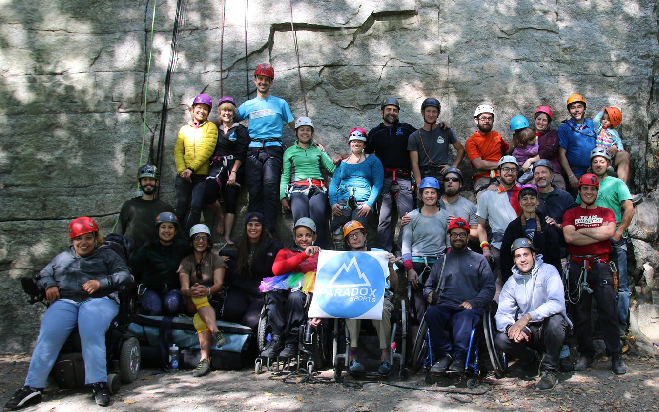 Group of people with and without disabilities posing for a photo in front of a rock wall holding the Paradox Sports Flag.
