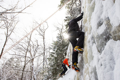Two ice climbers on a frozen waterfall with snow-covered trees in the background.