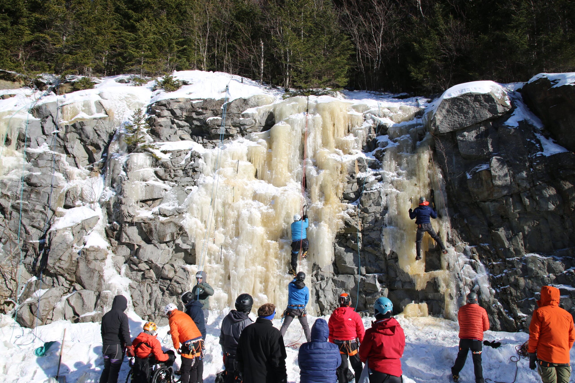 People ice climbing on a frozen waterfall in a snowy landscape.