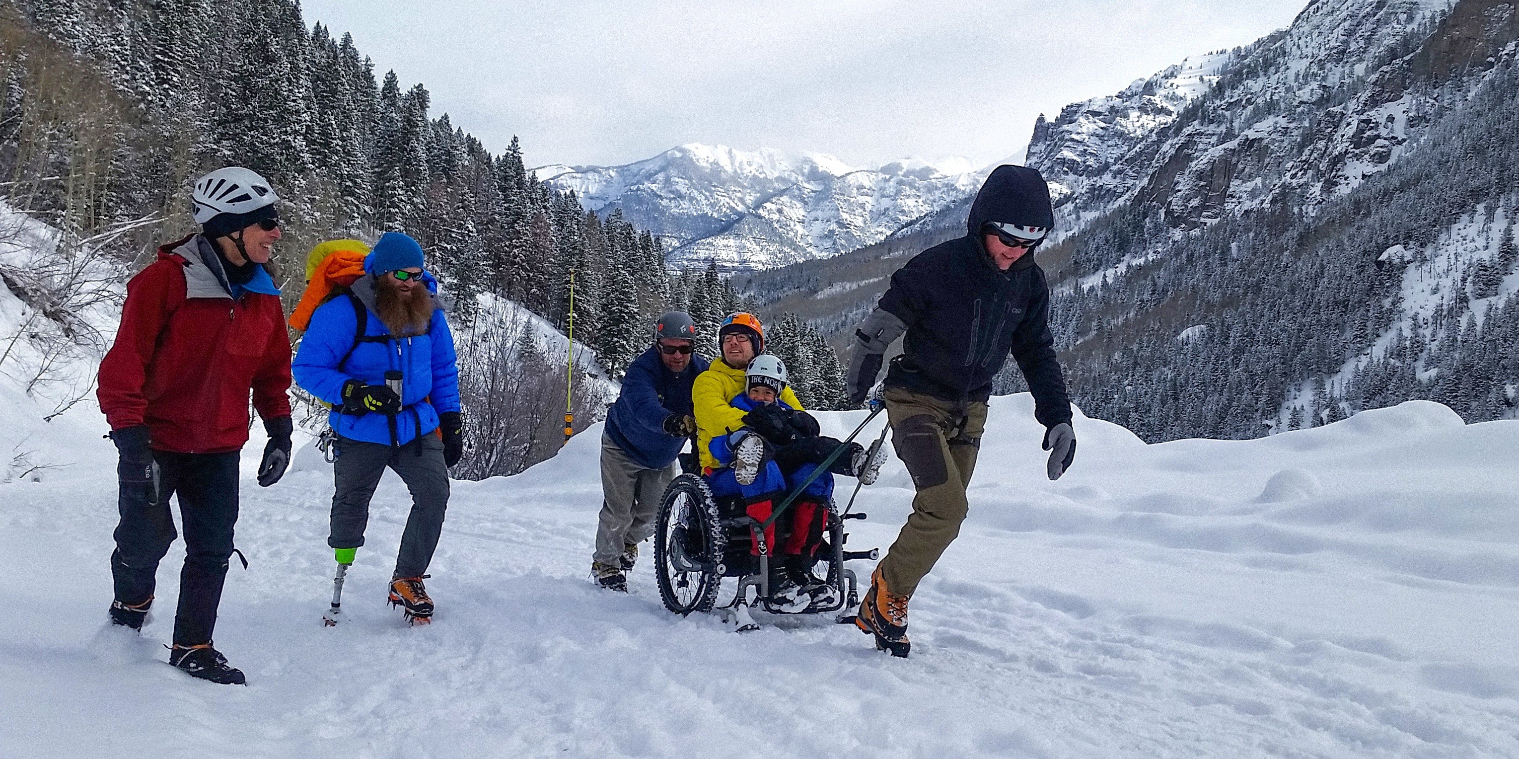 Group of people, including a person in a wheelchair, walking through a snowy landscape with mountains in the background.