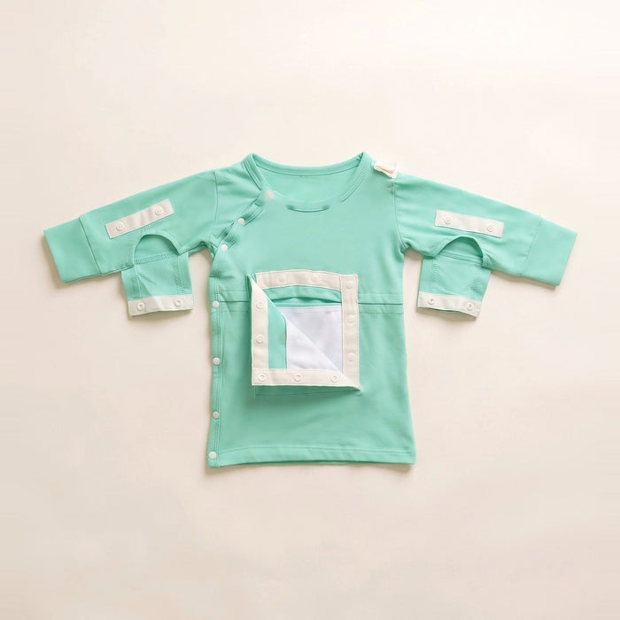 Front angle of the PediaTech Apparel LineWear Central Line Access Baby Gown in mint green (Calm Mint Edition) with the chest pocket flap opened to show its white lining and both sleeve openings unsnapped, laid flat against an off-white background