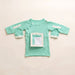 Front angle of the PediaTech Apparel LineWear Central Line Access Baby Gown in mint green (Calm Mint Edition) with the chest pocket flap opened to show its white lining and both sleeve openings unsnapped, laid flat against an off-white background