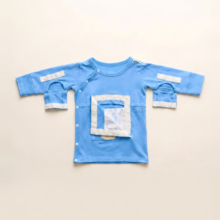 Front angle of the PediaTech Apparel LineWear Central Line Access Baby Gown in light blue (Sky Lullaby Edition) with the chest pocket flap opened to show its white lining and both sleeve openings unsnapped, laid flat against an off-white background