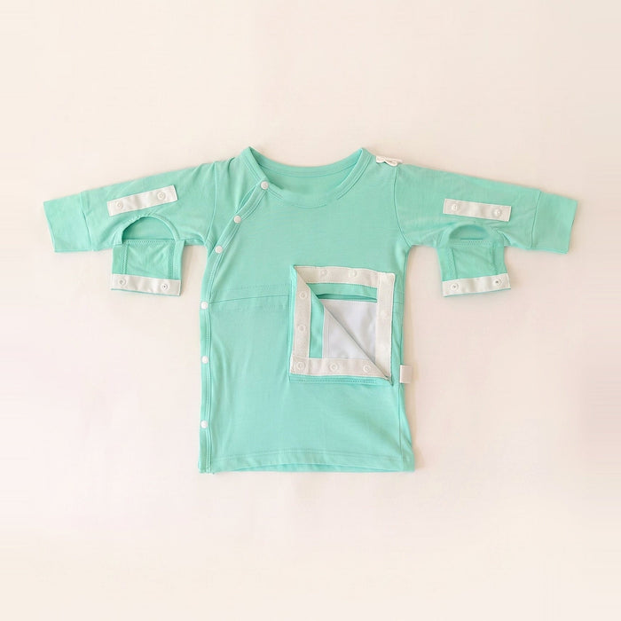 Front angle of the PediaTech Apparel TubeWear G/J Tube Access Baby Gown in mint green (Calm Mint Edition) with the abdomen pocket flap opened to show its white lining and both sleeve openings unsnapped, laid flat against an off-white background