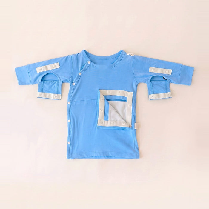 Front angle of the PediaTech Apparel TubeWear G/J Tube Access Baby Gown in light blue (Sky Lullaby Edition) with the abdomen pocket flap opened to show its white lining and both sleeve openings unsnapped, laid flat against an off-white background