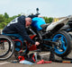 Person in a wheelchair working on a blue motorcycle with a clear sky background