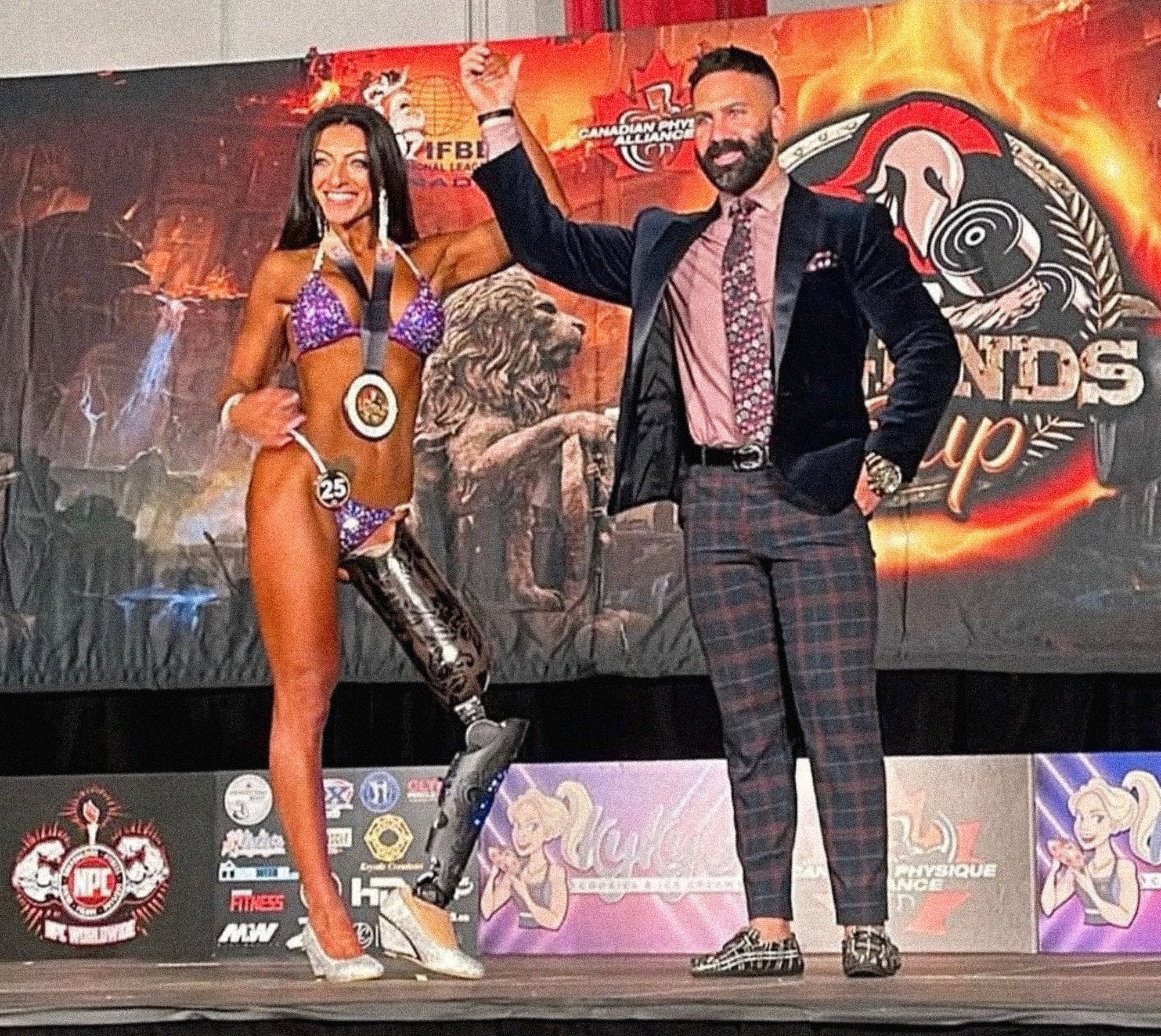 Rouzalin Hakim on the stage of a bodybuilding competition with her hand being raised by the event organizer in celebration of her placement in the competition