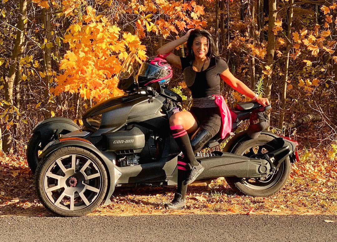 Portrait of Rouzalin Hakim sitting on the side of her 3 wheel Can-Am motorcycle in the autumn woods