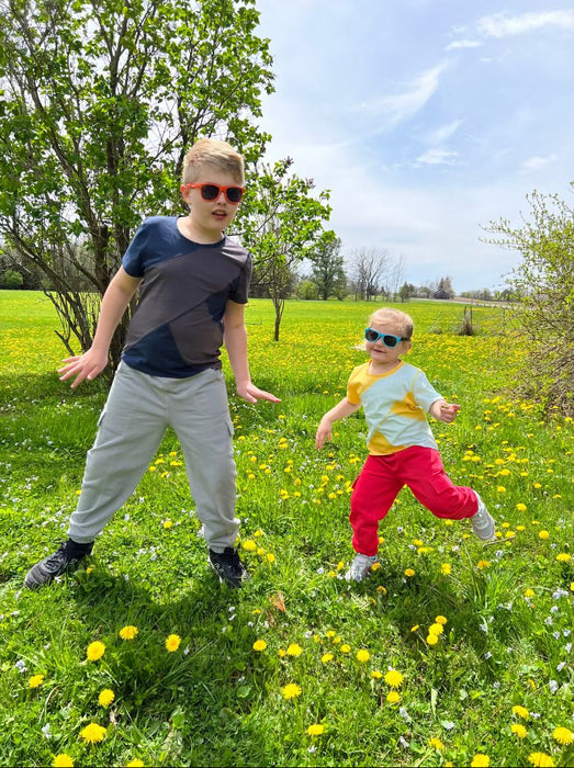 Two children playing in a grassy field with yellow flowers on a sunny day while wearing Sense-ational You Sensory Friendly Youth T-Shirts