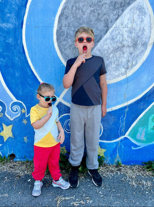 Two children posing in front of a colorful mural with sunglasses while wearing Sense-ational You Sensory Friendly Youth T-Shirts