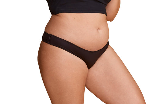 A close-up side view of a model wearing Slick Chicks Adaptive Tanga Panty in Black against a solid white background.