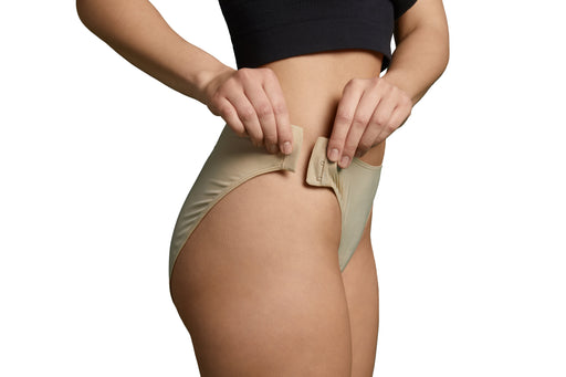 A close-up of a model wearing Slick Chicks Adaptive High Waist Brief in beige, pulling the hook & eye side closures closed with both hands, against a plain white background