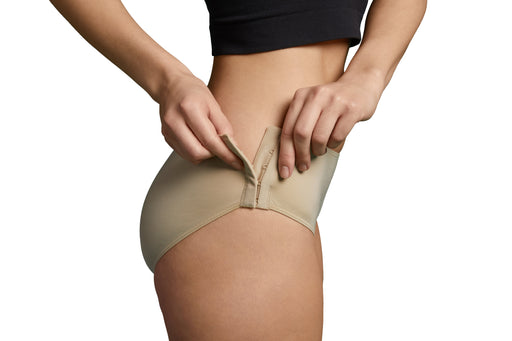 Close-up of a model wearing Slick Chicks Adaptive Hipster Panty in beige, pulling the hook & eye side closures with both hands against a plain white background