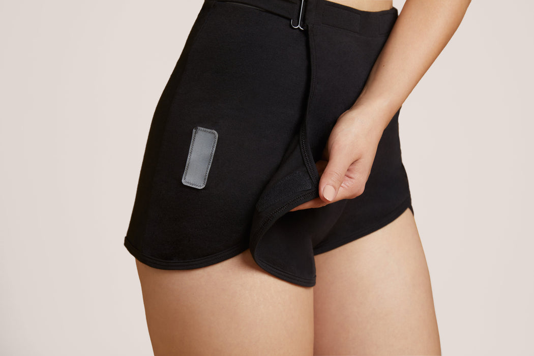 Close-up of a model wearing Slick Chicks Adaptive Lounge Short in black, demonstrating the VELCRO Brand Fasteners that detach the front panel, against a beige background