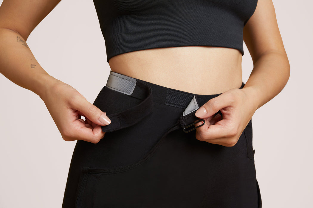 Close-up of a model wearing Slick Chicks Adaptive Lounge Short in black, demonstrating the VELCRO Brand Fasteners that detach the front panel along the ribbed waist band, against a beige background