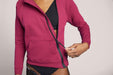 Close-up of Back angle of a model wearing the Slick Chicks Adaptive Magnetic Zip-Up Hoodie in plum, pulling the magnetic zipper open with one hand, against a beige background