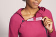 Close-up of a model wearing the Slick Chicks Adaptive Magnetic Zip-Up Hoodie in plum, pulling the collar open with one hand for port tube access, against a beige background
