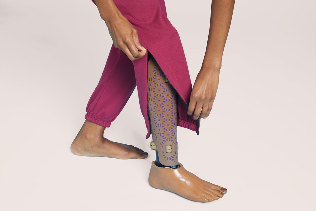 Close up of a model wearing Slick Chicks Adaptive Side Zip Joggers in plum, unzipping the side of the jogger which shows their prosthetic leg, against a plain beige background