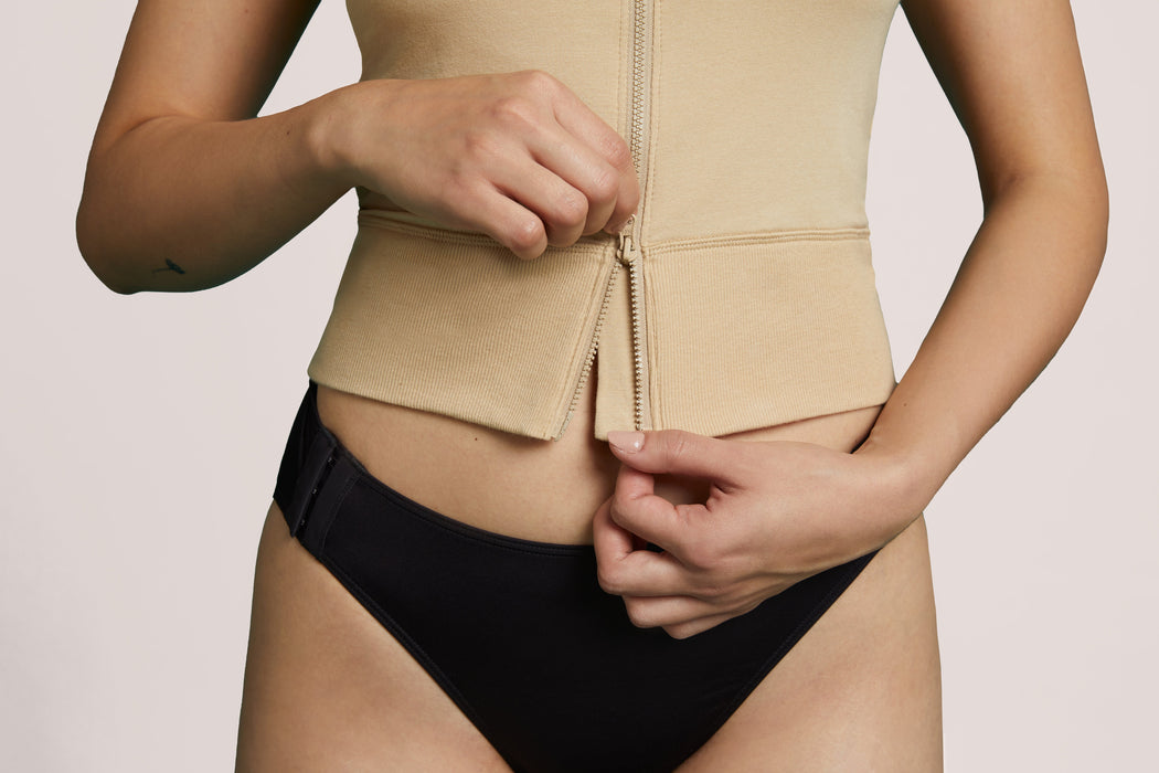 A close-up of a model wearing Slick Chicks Adaptive Two-Way Front Zip Cami in beige, demonstrating the two-way front zipper closure, against a plain beige background