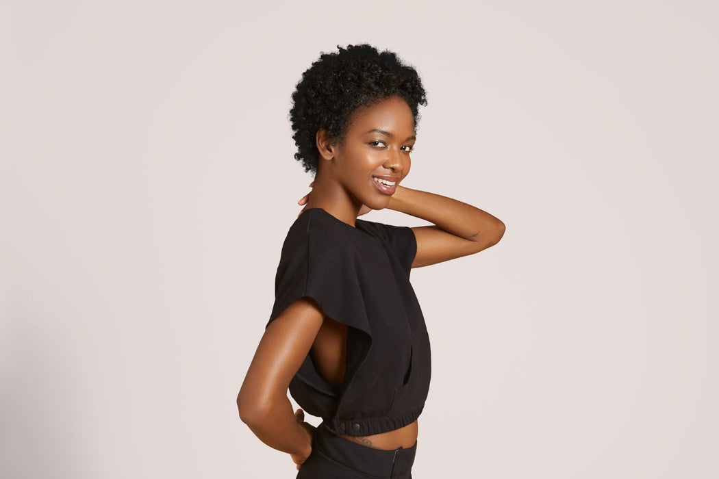 Side angle of a smiling woman with short curly black hair wearing Slick Chicks Adaptive V-Neck Crop Top in black, against a solid beige background