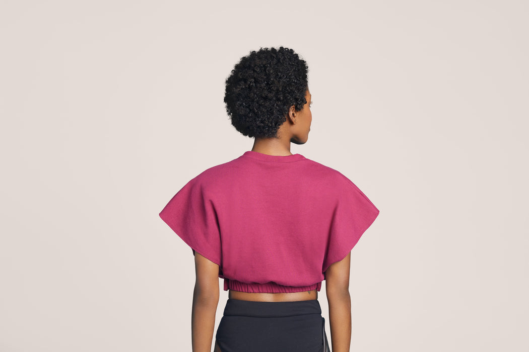Back angle of a woman with short curly black hair wearing Slick Chicks Adaptive V-Neck Crop Top in plum, against a solid beige background