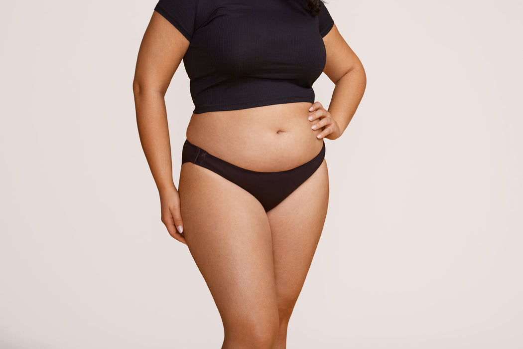 Front angle of a model wearing Slick Chicks Adaptive Bikini Brief Panty in black against a plain light beige studio background