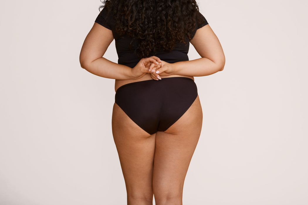 Back angle of a model wearing Slick Chicks Adaptive Bikini Brief Panty in black against a plain light beige studio background