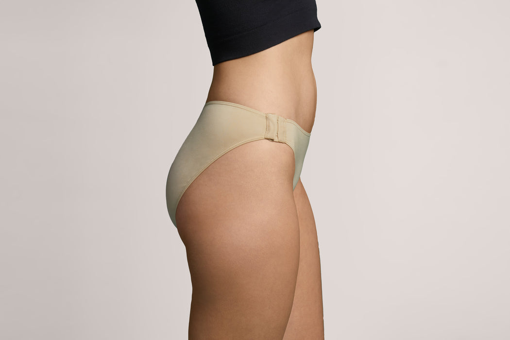 Side angle of a model wearing Slick Chicks Adaptive Bikini Brief Panty in beige against a plain light beige studio background
