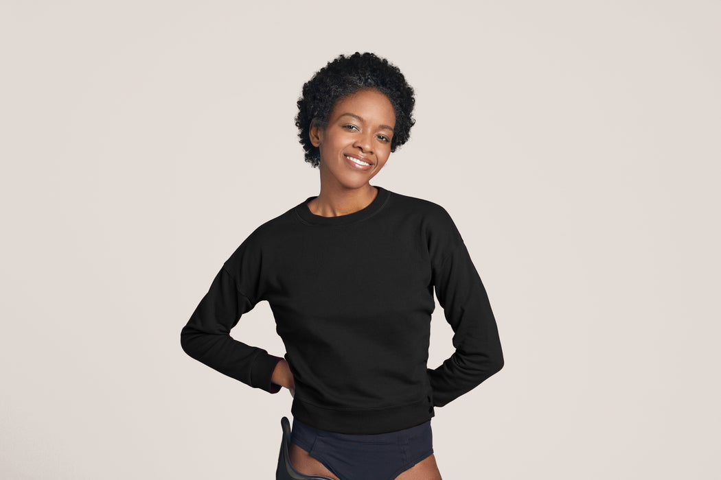 Front angle of a smiling woman with short black curly hair wearing Slick Chicks Adaptive Crewneck Sweatshirt in black, against a solid beige background