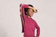 A smiling woman with short black curly hair wearing Slick Chicks Adaptive Crewneck Sweatshirt in plum, resting her arm on her forehead demonstrating the VELCRO Brand Fasteners along the side of the Sweatshirt against a solid beige background