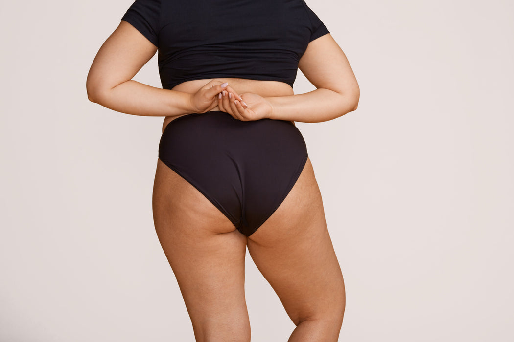 Rear angle of a model wearing Slick Chicks Adaptive High Waist Brief in black against a plain light beige studio background