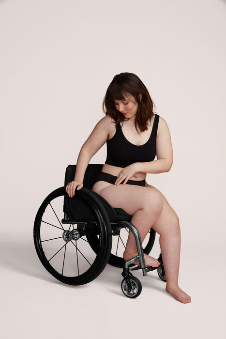 A model wearing Slick Chicks Adaptive High Waist Brief in black, seated on a black manual wheelchair against a beige white background